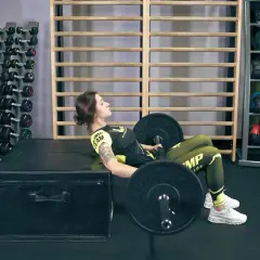 Kobieta wykonuje hip thrust po polsku, z ciężarem na biodrach, opierając górną część pleców na skrzyni.