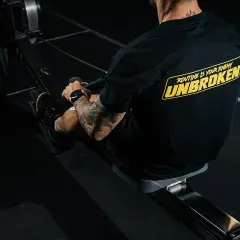 Mężczyzna z tatuażami na ramieniu trenuje na wioślarzu. Czy to crossfit czy siłownia, ważne, że jest UNBROKEN!