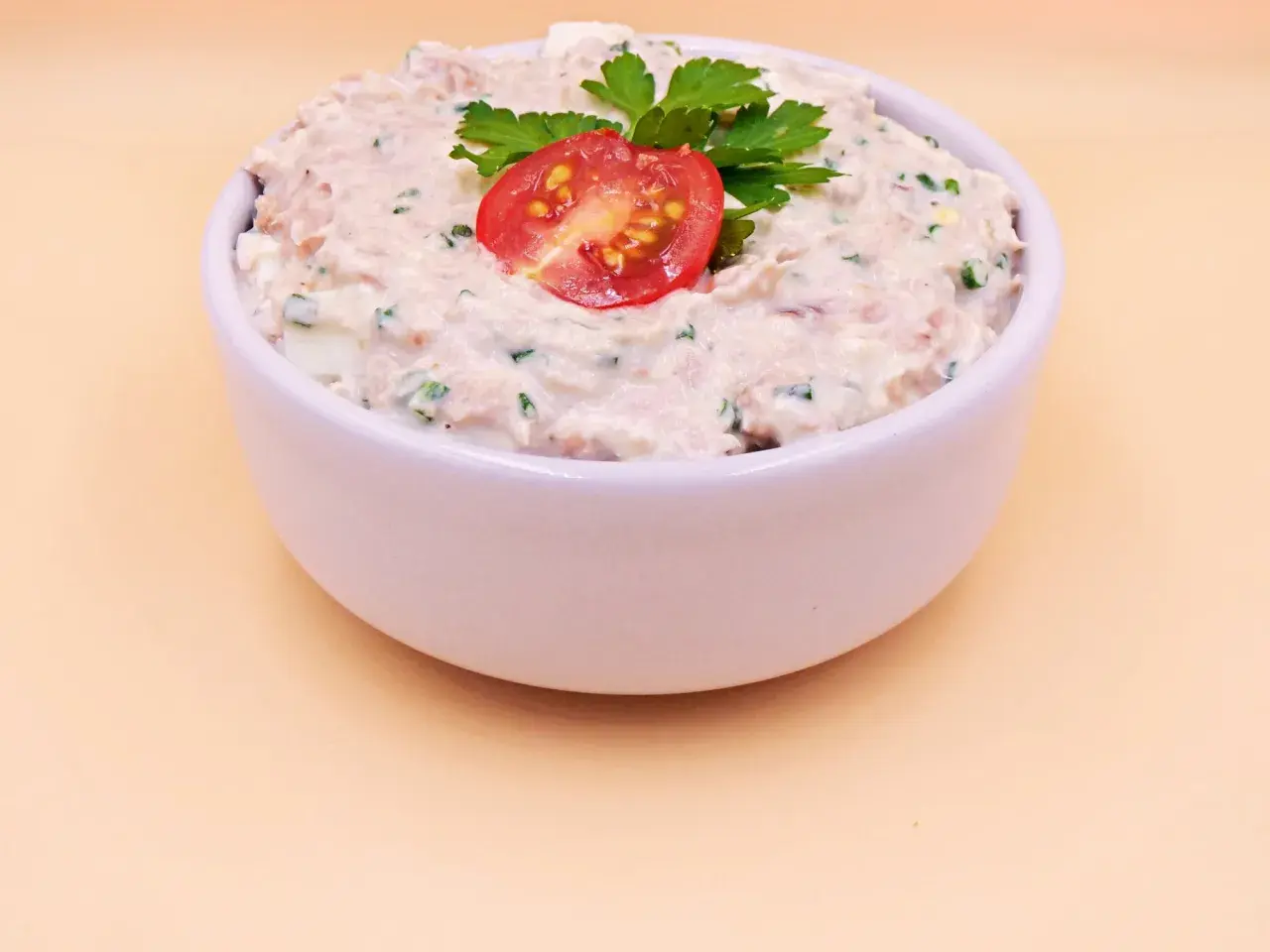 Pyszna pasta z tuńczyka i jajka, udekorowana pomidorkiem i natką pietruszki, w białej miseczce.