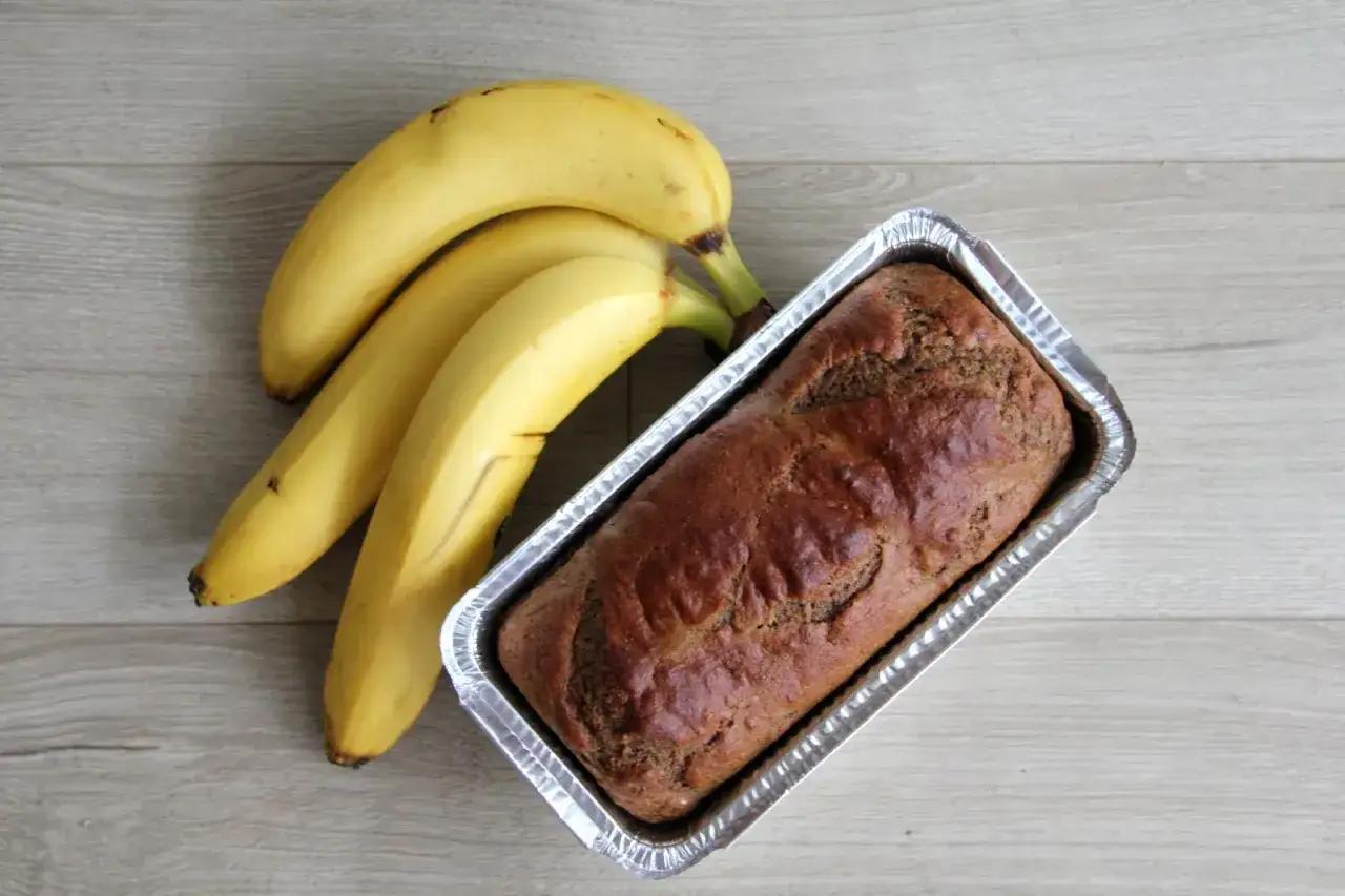 Gotowy proteinowy chlebek bananowy w formie, obok świeże banany na drewnianym tle.