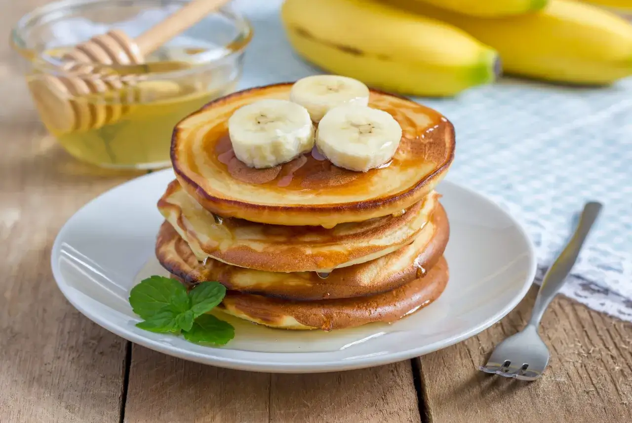 Stos placuszków bananowych polanych miodem, ozdobiony plasterkami banana i listkiem mięty. Obok miseczka z miodem i widelczyk.